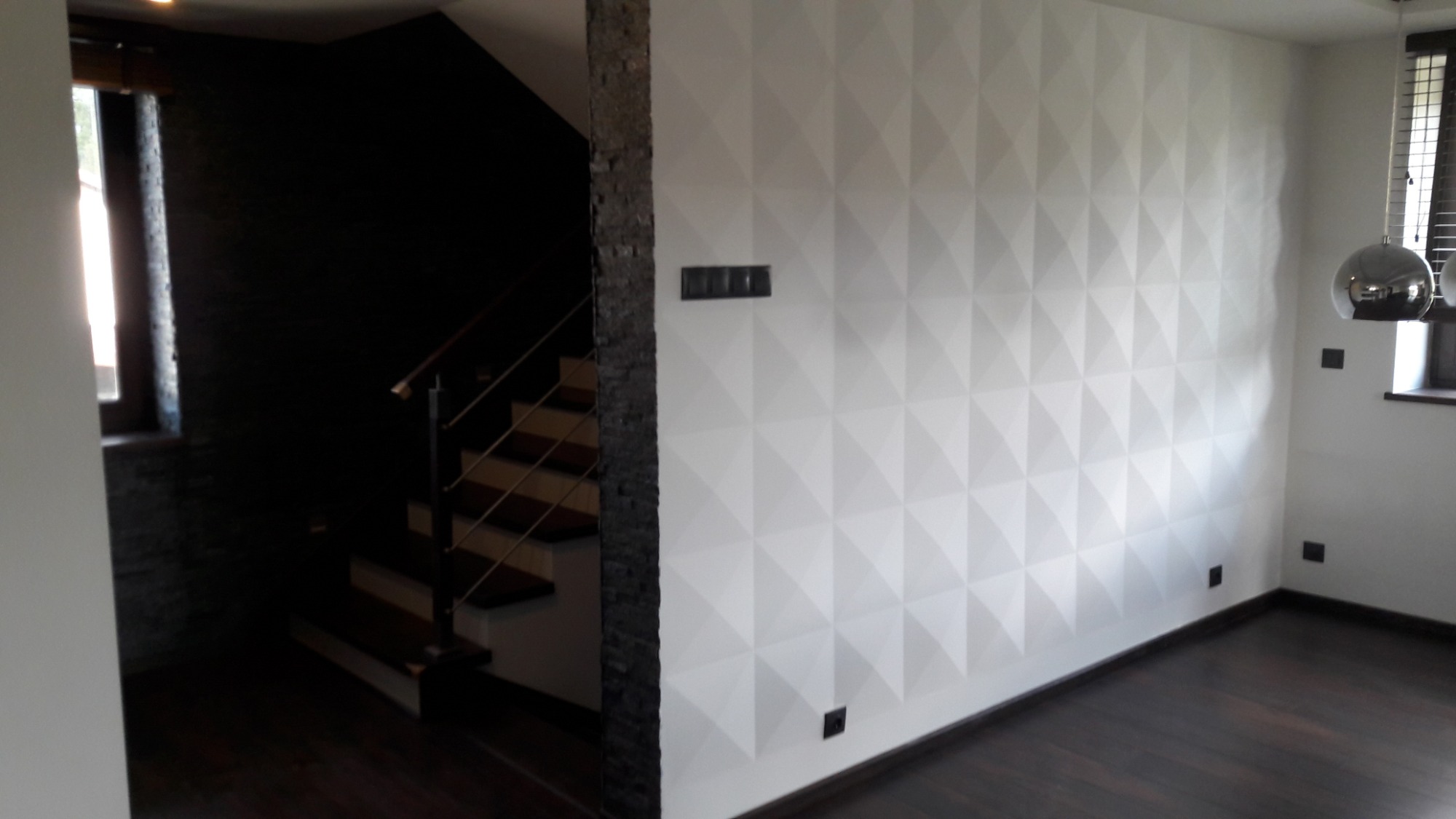 Biała ściana z trójwymiarowym wzorem piramid, ciemne drewniane schody z metalową balustradą i czarna ściana w tle, ciemna podłoga, nowoczesne oświetlenie wiszące.