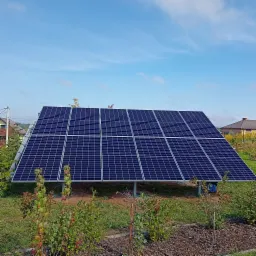 Panel fotowoltaiczny ustawiony na metalowej konstrukcji na zewnątrz, w tle domy jednorodzinne i błękitne niebo z delikatnymi chmurami.