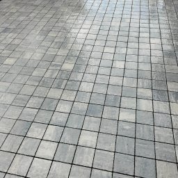 Artur - Ułożona, szara kostka brukowa w kwadraty z czarnymi fugami, lekko mokra powierzchnia odbija niebo.