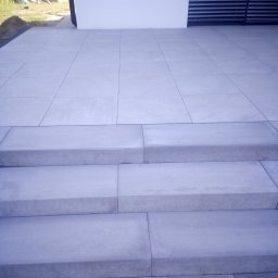 Artur - Nowoczesne schody wejściowe i taras wyłożone dużymi, szarymi płytami betonowymi, prowadzące do budynku z białymi ścianami i żaluzjami w oknach.