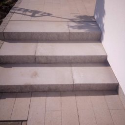 Artur - Jasne, granitowe schody wejściowe prowadzące do budynku, z widocznym fragmentem chodnika z kostki i cieniem rzucanym przez drzewo.