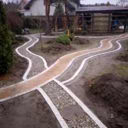 Kręte ścieżki w ogrodzie: jedna z kostki w odcieniach beżu, druga wysypana szarym żwirem, obie obramowane jasnymi obrzeżami, prowadzące przez świeżo przekopaną ziemię z widocznymi drzewami...