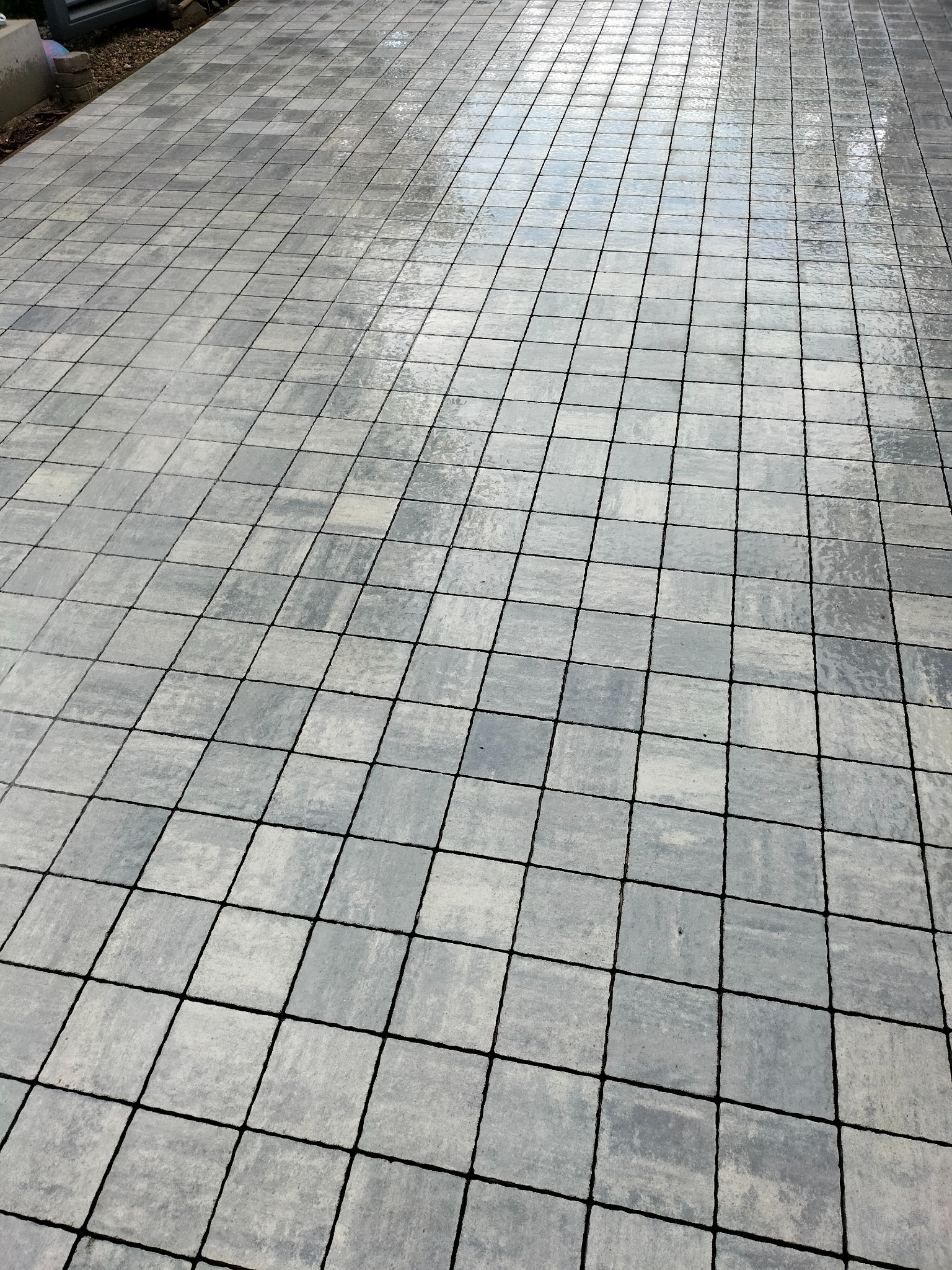Ułożona, szara kostka brukowa w kwadraty z czarnymi fugami, lekko mokra powierzchnia odbija niebo.