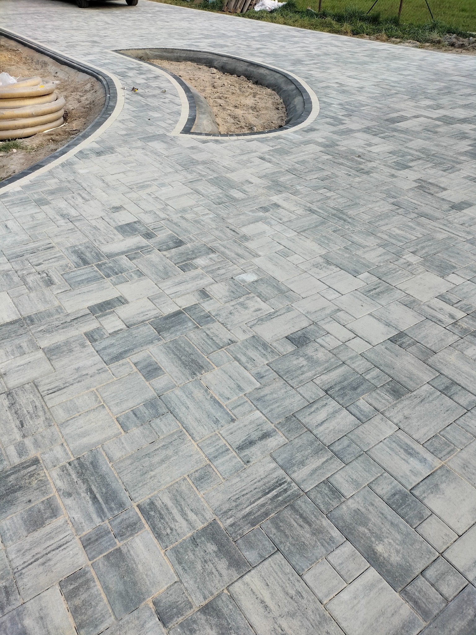 Ułożona nawierzchnia z szaro-białej kostki brukowej o nieregularnym wzorze, z obrzeżem wykończonym ciemniejszymi elementami, obok leżą rury karbowane, w tle trawnik.