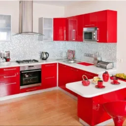 Nowoczesna kuchnia z błyszczącymi, czerwonymi frontami szafek, mozaiką na ścianie i drewnianą podłogą. Na blacie ekspres do kawy, czajnik i toster.