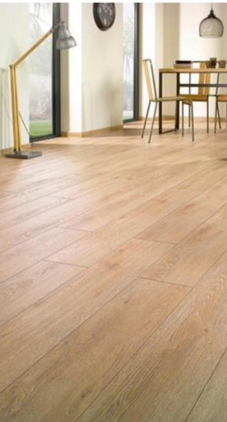 Jasna, drewniana podłoga ułożona w jodełkę, widoczna lampa podłogowa z metalowym kloszem, okno i fragment stołu z krzesłami w tle.