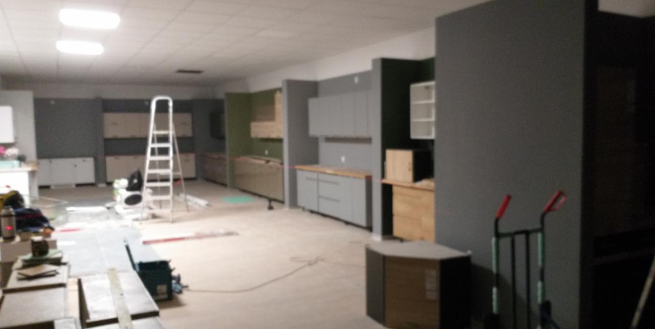 Wnętrze w trakcie montażu zabudowy kuchennej; widoczne surowe szafki w różnych kolorach, drabina oraz materiały budowlane.