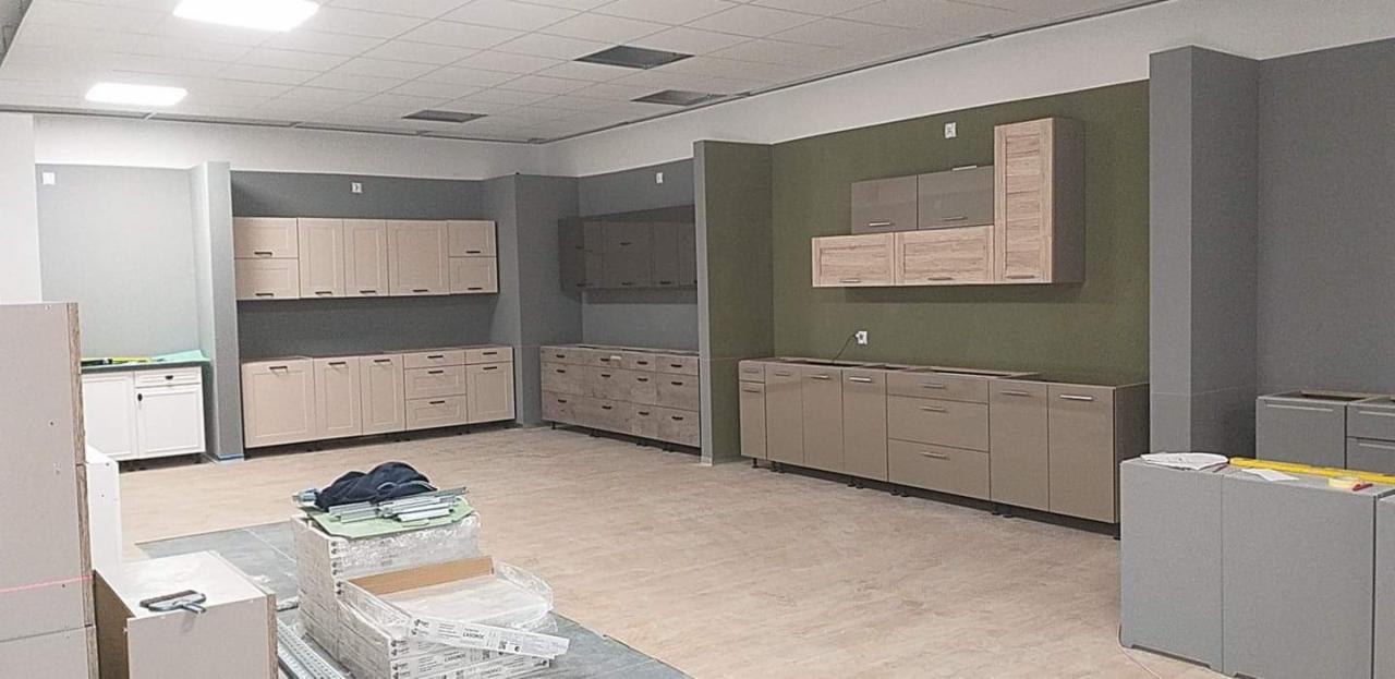 Wnętrze w trakcie montażu zabudowy kuchennej z beżowymi frontami, widoczne częściowo rozpakowane elementy i narzędzia na podłodze, ściany w odcieniach szarości i oliwkowej zieleni.