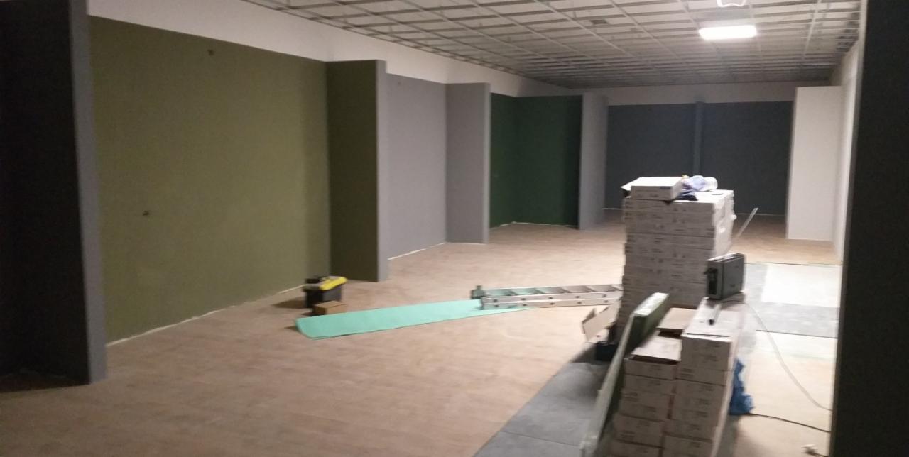 Wnętrze w trakcie prac malarskich: pomalowane ściany w odcieniach zieleni i szarości, na podłodze rozłożona folia ochronna i drabina, w tle stos kartonów z płytkami.