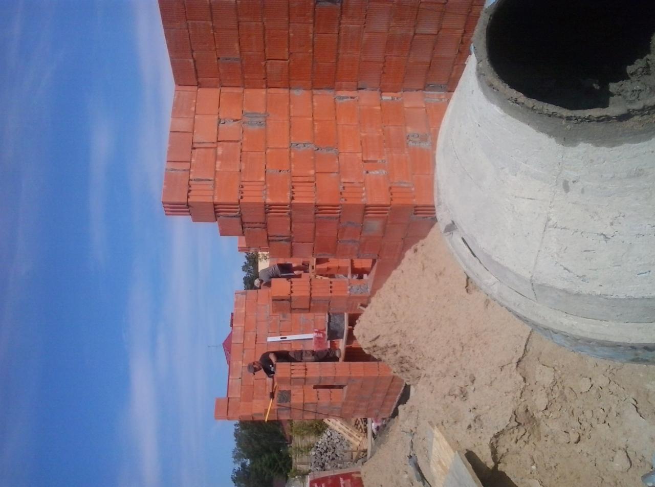 Wznoszenie ścian z czerwonej cegły, widoczna betoniarka i dwóch murarzy w trakcie pracy. Budowa domu w słoneczny dzień.