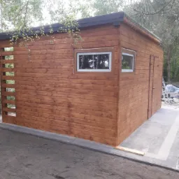 Nowoczesna, drewniana konstrukcja z dwoma oknami z białymi ramami i drzwiami, z elementami ażurowymi, pomalowana na brązowo, otoczona zielenią drzew.