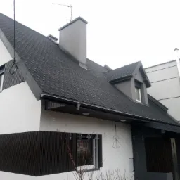 Nowoczesny dom z dachem pokrytym czarną dachówką bitumiczną, widoczne obróbki blacharskie w kolorze szarym, komin i antena na dachu.