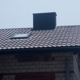 Ciemna blachodachówka z oknem dachowym i kominem na murowanym domu z cegły, widoczna rynna.