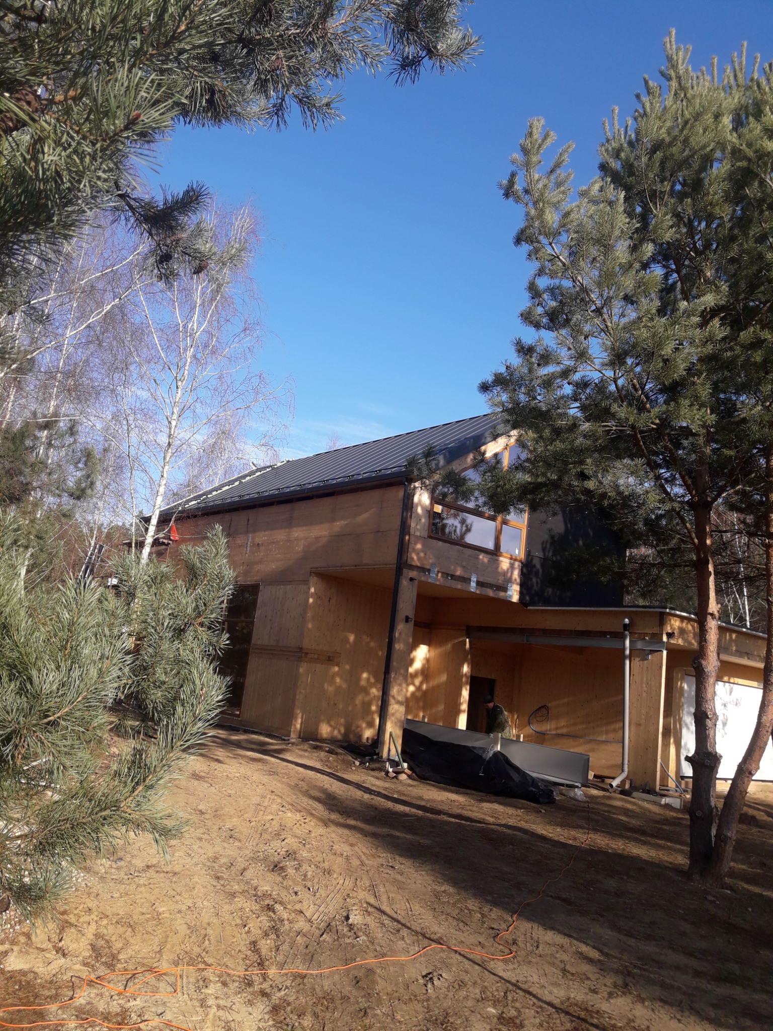 Wznoszony nowoczesny budynek z drewnianą elewacją i ciemnym dachem, widoczny w otoczeniu drzew iglastych i ziemistego terenu.