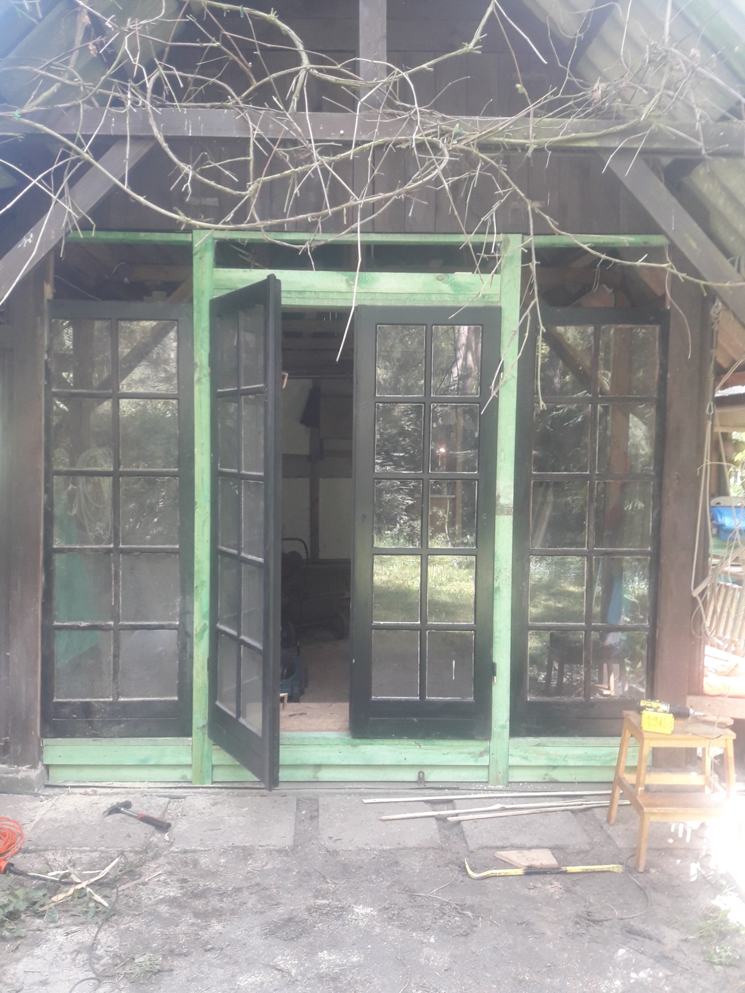 Drewniana altana w trakcie budowy z otwartymi czarnymi drzwiami z szybami, widoczne narzędzia pracy i drabinka.