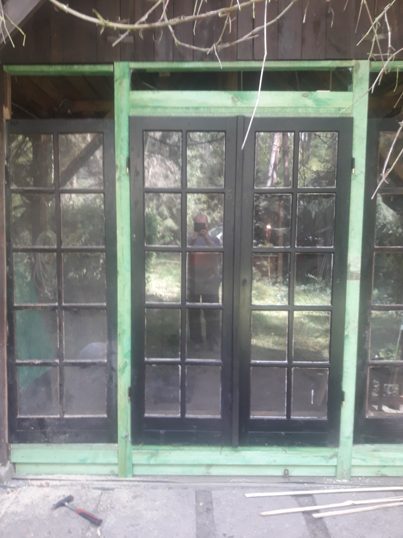 Trzyczęściowe okno z czarnymi ramami i zielonymi elementami konstrukcyjnymi, młotek i listwy na posadzce, widoczne odbicie osoby w szybie.