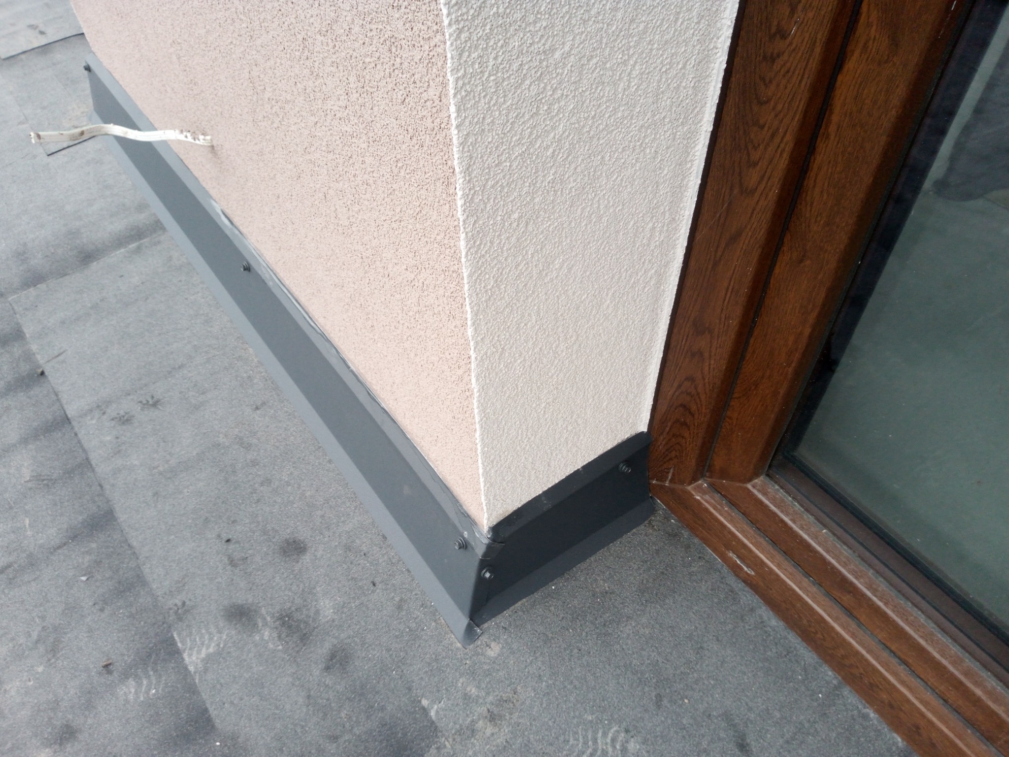Szczegółowe ujęcie narożnika dachu z obróbką blacharską w kolorze grafitowym, przylegającego do ściany z jasnym tynkiem strukturalnym i brązowej ramy okiennej. Widoczny fragment kabla wystający ze...