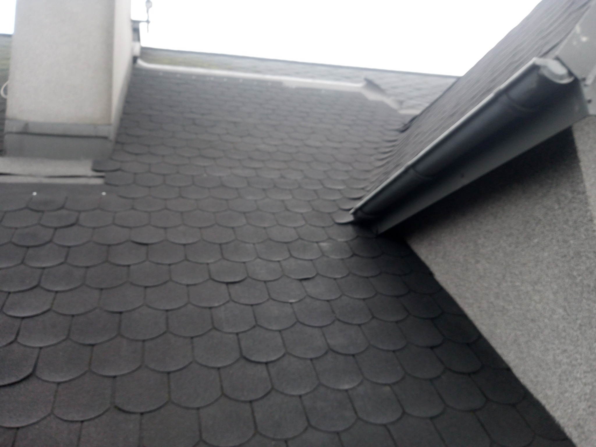 Dach pokryty gontem bitumicznym w kształcie rybiej łuski, widoczny komin i fragment rynny.