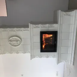 Biały, kaflowy piec kominkowy z paleniskiem, ozdobiony płaskorzeźbą z motywem zwierzęcym, w surowym wnętrzu.