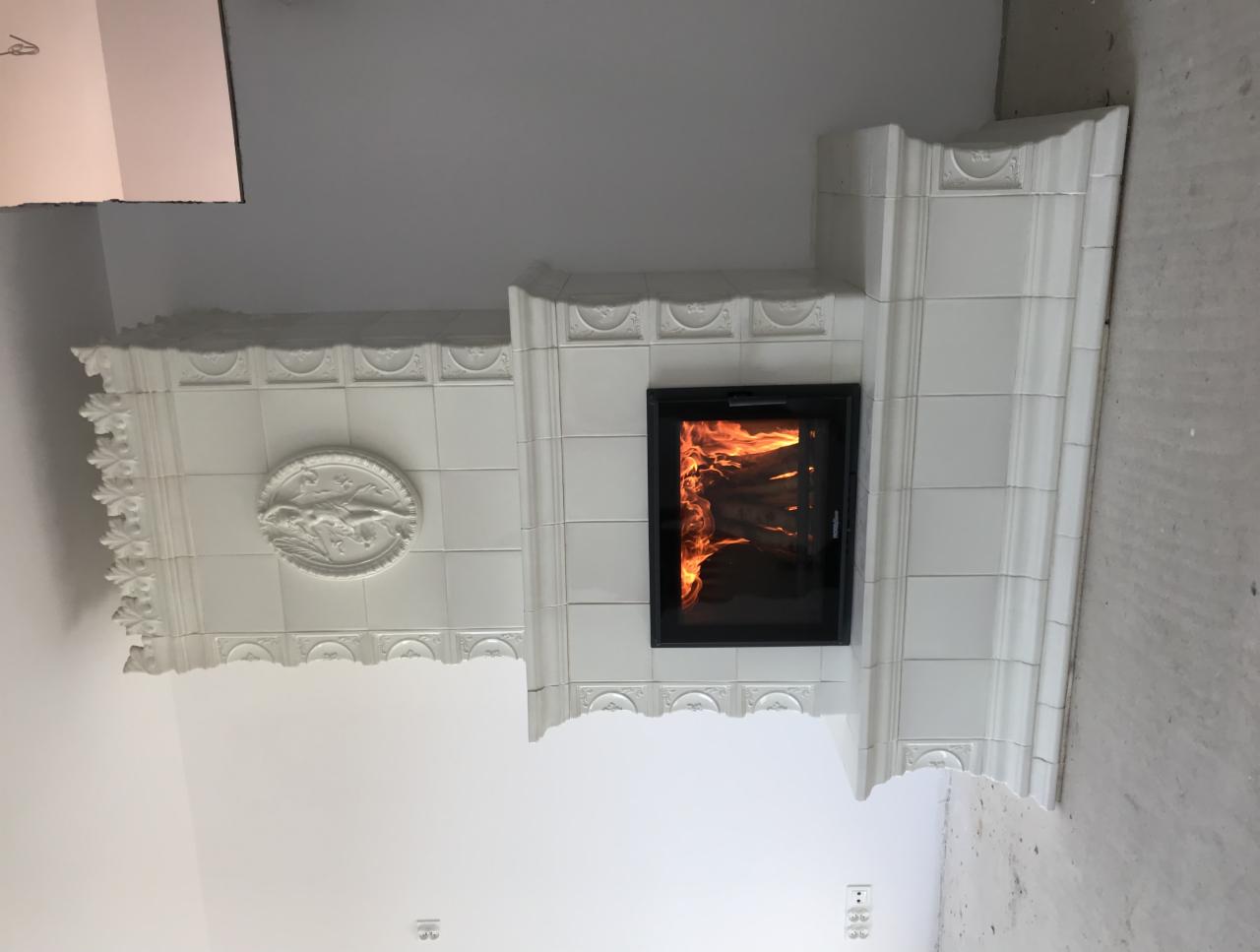 Biały, kaflowy piec kominkowy z paleniskiem, ozdobiony płaskorzeźbą z motywem zwierzęcym, w surowym wnętrzu.