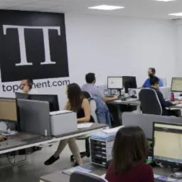 Jasne biuro z pracownikami przy biurkach z komputerami, logo firmy na ścianie. Oświetlenie sufitowe. Widok na przestrzeń biurową.