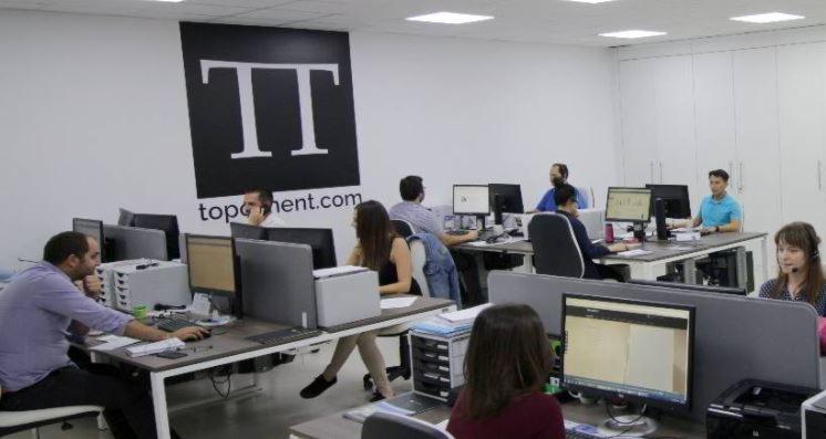 Jasne biuro z pracownikami przy biurkach z komputerami, logo firmy na ścianie. Oświetlenie sufitowe. Widok na przestrzeń biurową.