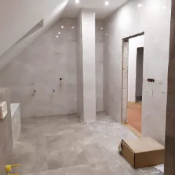 Wnętrze łazienki w trakcie remontu w domu ze skośnym dachem, widoczne płytki podłogowe i ścienne w odcieniach szarości, niezabudowana wanna, instalacja hydrauliczna, karton na podłodze...