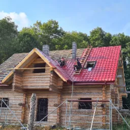Budowa drewnianego domu z czerwonym dachem z dachówki, widoczni dwaj pracownicy na dachu, jeden siedzi na drabinie, drugi obok komina, w tle las i błękitne niebo.