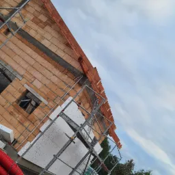 Budowa domu jednorodzinnego w trakcie ocieplania ścian styropianem, widoczne rusztowania, cegły, elementy konstrukcji dachu oraz niebieskie rury wentylacyjne na ziemi.