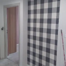 Ściana w kratę w odcieniach szarości i bieli w mieszkaniu w Krakowie, widoczny fragment drzwi i podłoga z szarych płytek, w rogu oparta o ścianę różowa rączka wałka malarskiego.