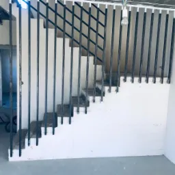 Wnętrze w trakcie remontu z surowymi schodami i zamontowanymi pionowymi, metalowymi elementami konstrukcji balustrady, widoczne kable i żarówka