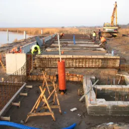 Budowa fundamentów nad brzegiem jeziora, widoczne zbrojenia, betonowe szalunki, robotnicy w odblaskowych ubraniach i koparka Hyundai w tle.