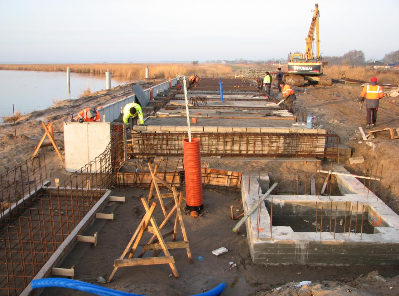 Budowa fundamentów nad brzegiem jeziora, widoczne zbrojenia, betonowe szalunki, robotnicy w odblaskowych ubraniach i koparka Hyundai w tle.
