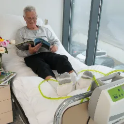 Pacjent w łóżku szpitalnym z aparatem do masażu pneumatycznego stóp, czytający magazyn, z bukietem kwiatów i jabłkiem na szafce nocnej.