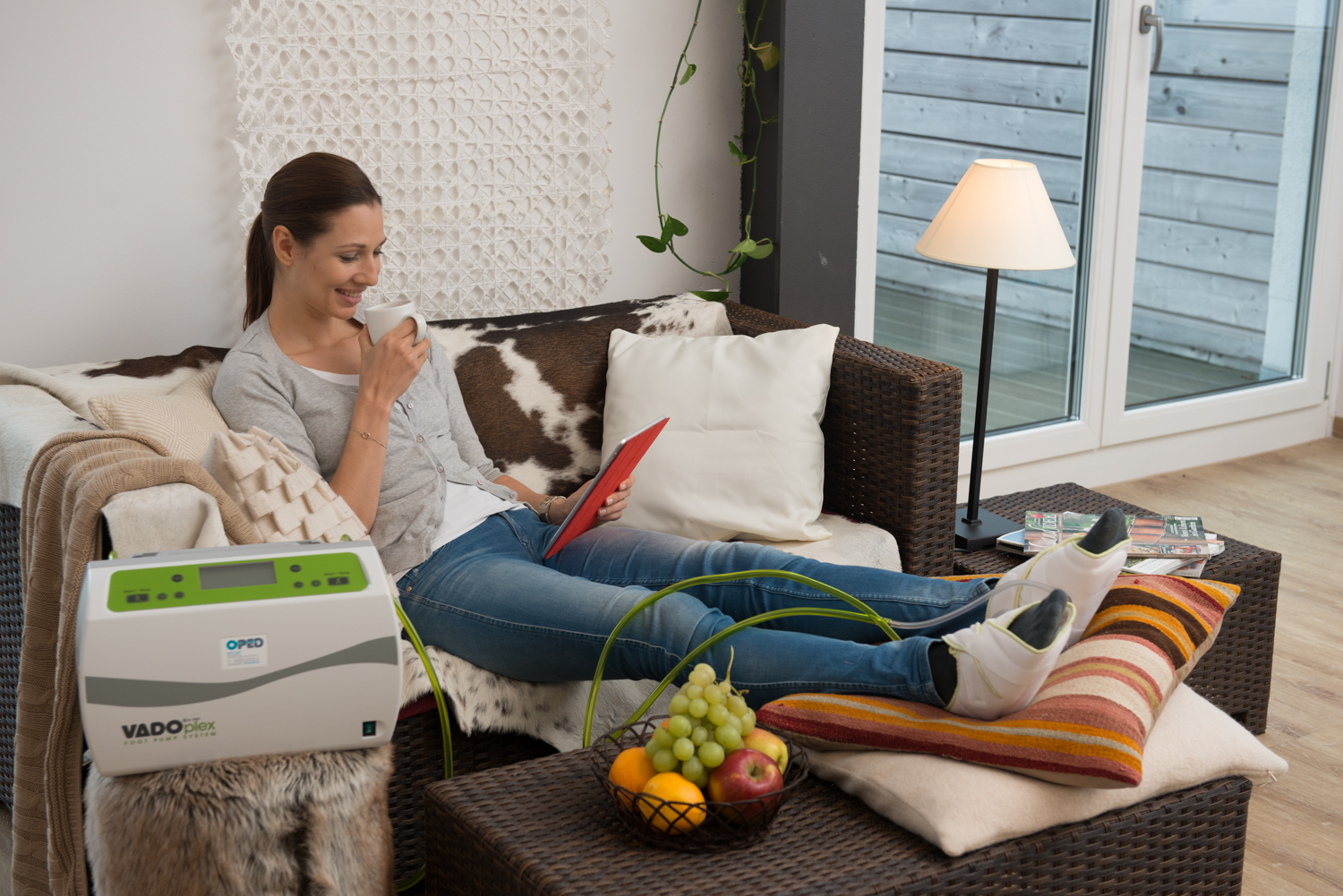 Kobieta w dżinsach relaksuje się na kanapie z kubkiem, używając urządzenia do masażu limfatycznego VADOPlex Oped, z nogami w specjalnych butach, czytając tablet i jedząc owoce.
