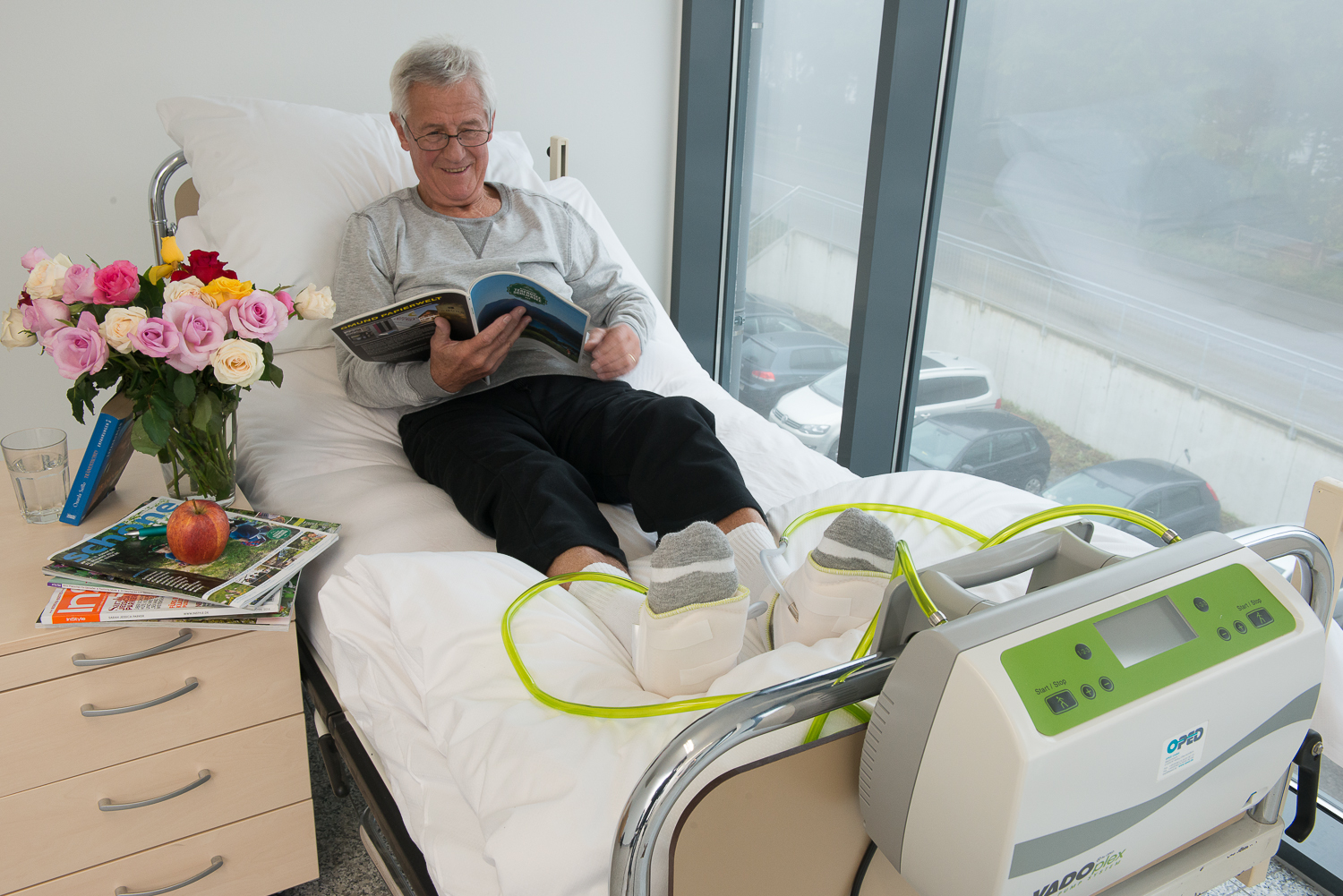 Pacjent w łóżku szpitalnym z aparatem do masażu pneumatycznego stóp, czytający magazyn, z bukietem kwiatów i jabłkiem na szafce nocnej.