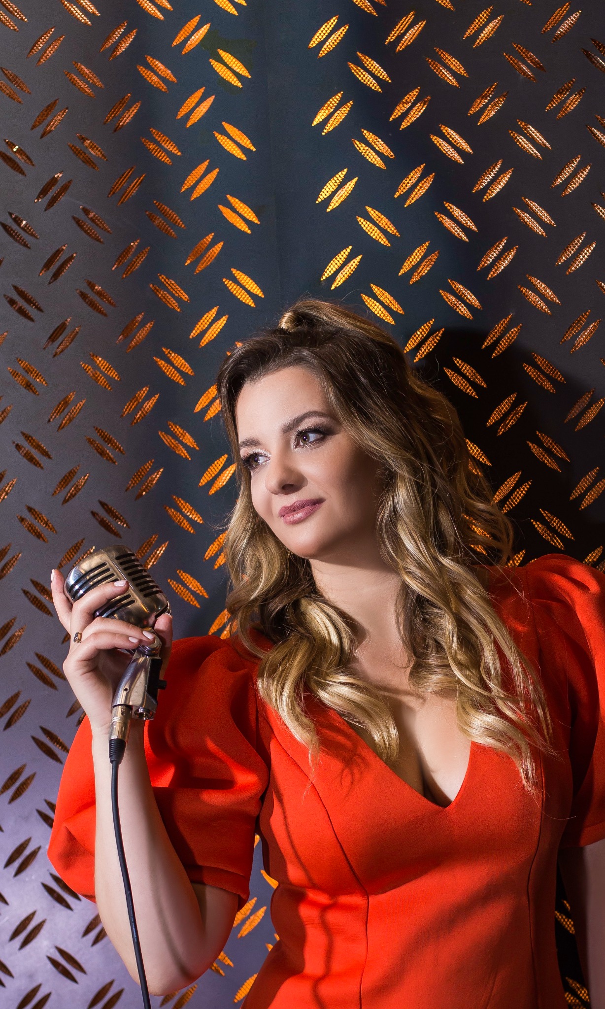 Kobieta w czerwonej sukience trzymająca srebrny mikrofon w stylu retro na tle metalowej ściany z pomarańczowymi akcentami.