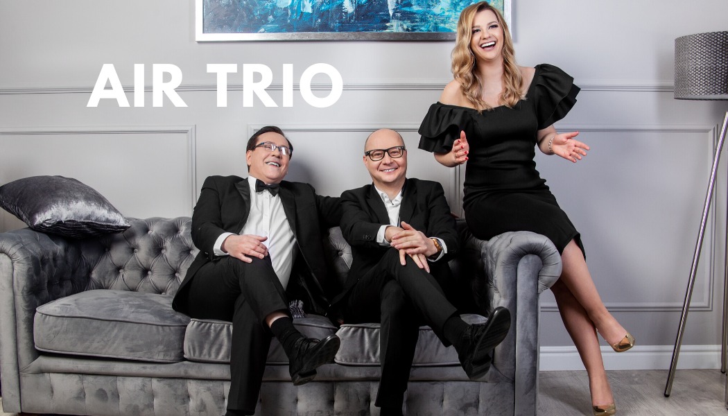 Promocyjne zdjęcie zespołu Air Trio: dwóch mężczyzn w smokingach siedzi na szarej sofie, a kobieta w czarnej sukience siedzi na oparciu sofy, wszyscy uśmiechnięci.