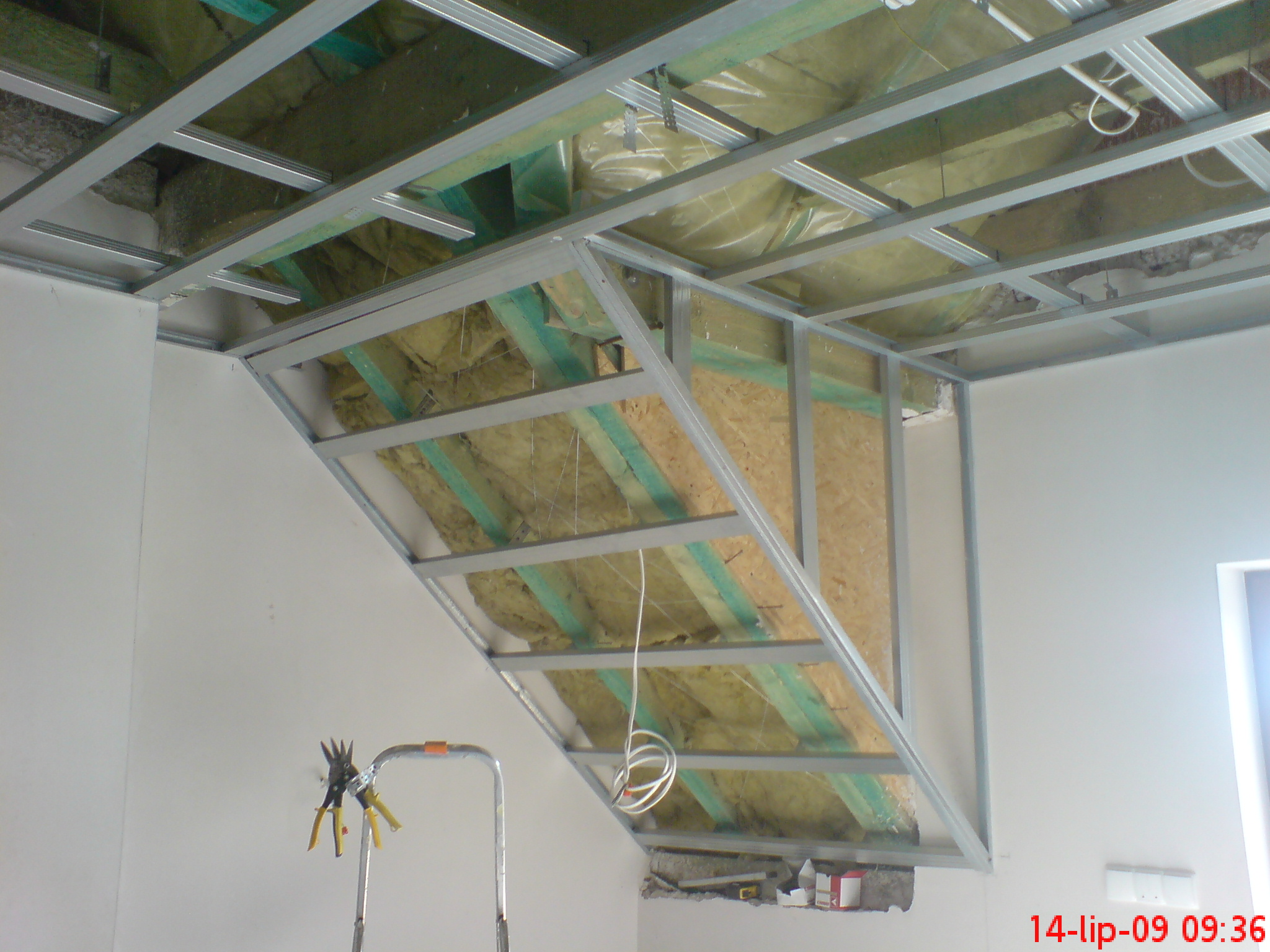 Szkielet metalowy pod zabudowę poddasza z izolacją z wełny mineralnej, widoczny fragment drabiny i narzędzia do cięcia profili.