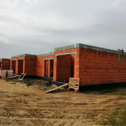 Surowy stan budowy szeregu domów z czerwonej cegły, widoczne otwory okienne i drzwiowe, paleta z cegłami i drewniane rusztowanie, ziemisty teren budowy pod pochmurnym niebem.