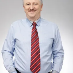 Uśmiechnięty mężczyzna z wąsami, ubrany w błękitną koszulę w paski i czerwono-biało-granatowy krawat, pozuje z rękami w kieszeniach spodni.