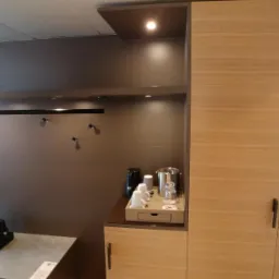 Nowoczesna szafa z jasnego drewna z wbudowaną p&oacute;łką i oświetleniem, widoczny zestaw do parzenia kawy i ł&oacute;żko w tle.