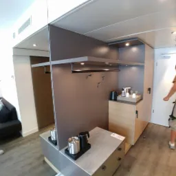 Wnętrze kabiny na statku wycieczkowym z zabudową meblową: szafa z wieszakami, półka z oświetleniem LED, szafka z czajnikiem i zestawem do kawy/herbaty, oraz komoda z akcesoriami.