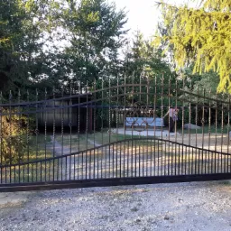 Czarna, metalowa brama wjazdowa z ozdobnymi elementami w Włocławku, widoczna z perspektywy podwórza, z zarysem osoby i samochodu w tle.