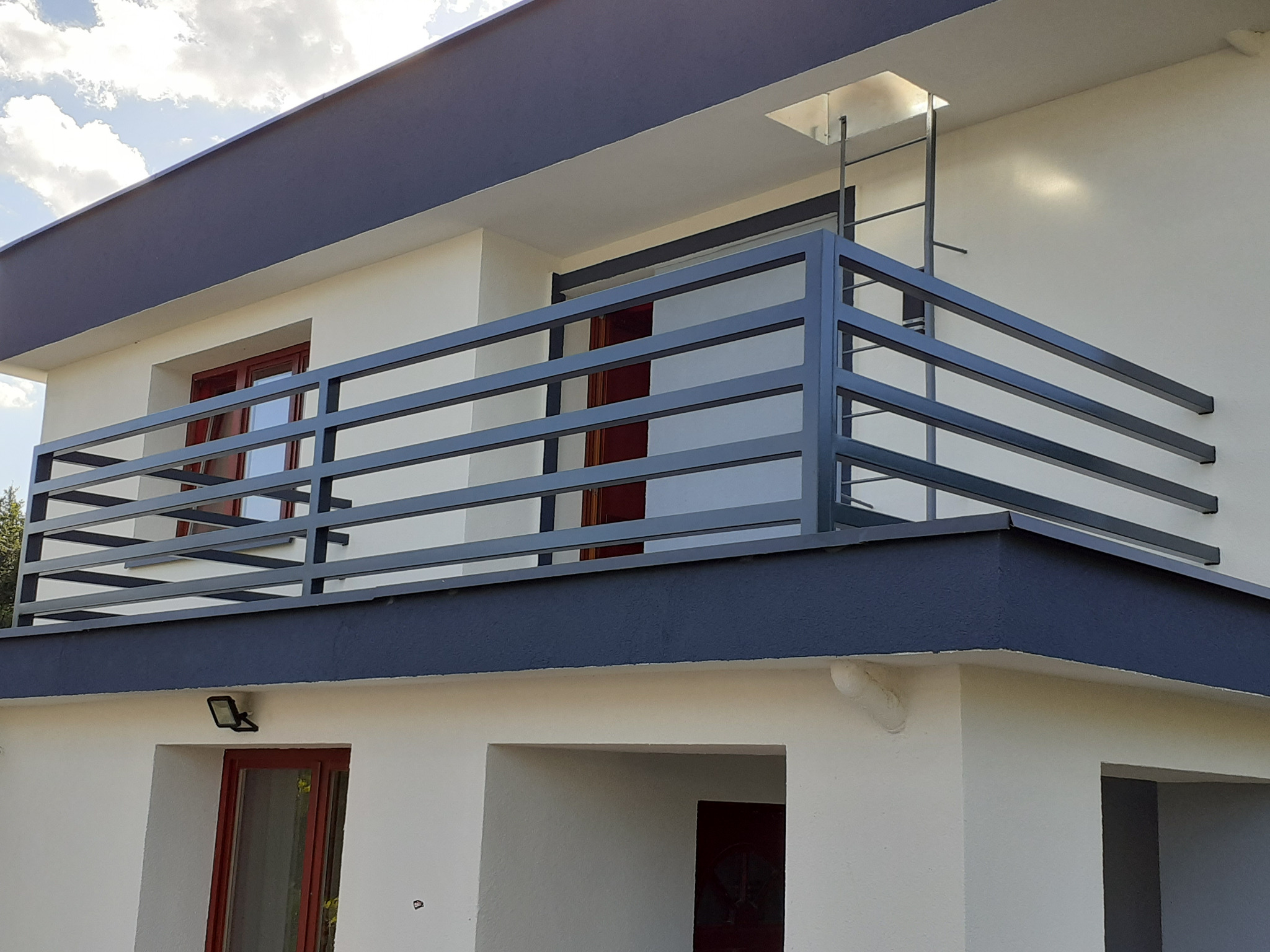 Nowoczesna, szara balustrada balkonowa z poziomych profili na tle białej elewacji budynku z ciemnoszarym dachem i czerwonymi ramami okiennymi.