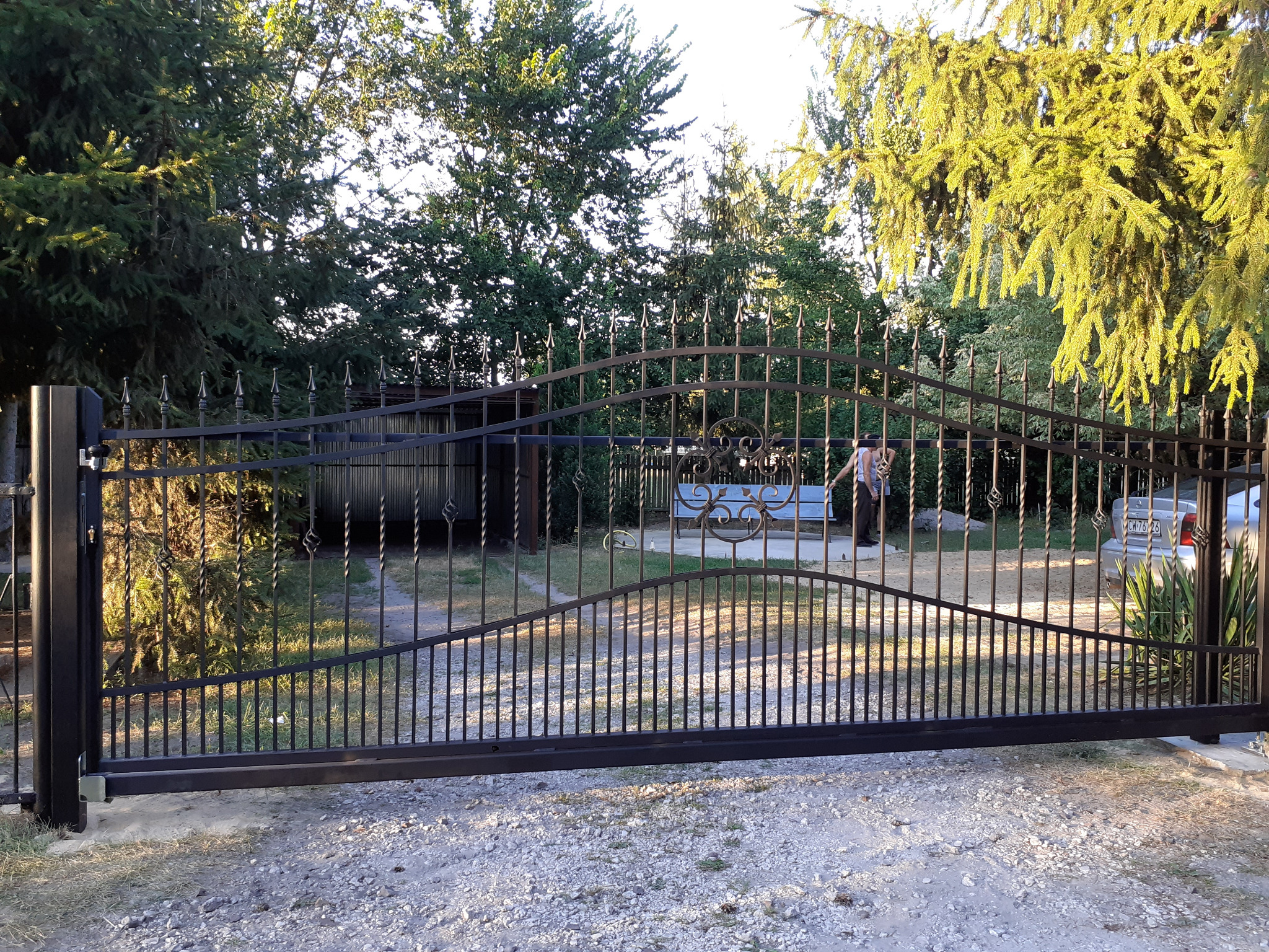 Czarna, metalowa brama wjazdowa z ozdobnymi elementami w Włocławku, widoczna z perspektywy podwórza, z zarysem osoby i samochodu w tle.