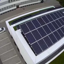 Widok z góry na budynek z panelami słonecznymi na dachu, obok parking z zaparkowanymi samochodami i zieleń.