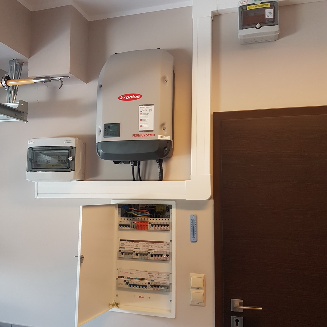 Zamontowany inwerter Fronius Symo obok otwartej skrzynki z bezpiecznikami, dodatkowo widoczny wyłącznik i termometr pokojowy.