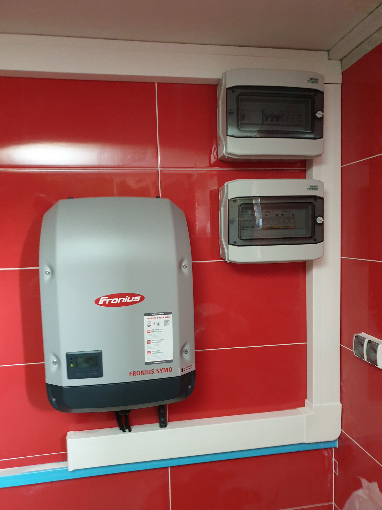 Szary inwerter Fronius Symo zamontowany na czerwonej, błyszczącej ścianie z płytek, obok dwie szare skrzynki z bezpiecznikami, biała listwa przypodłogowa.