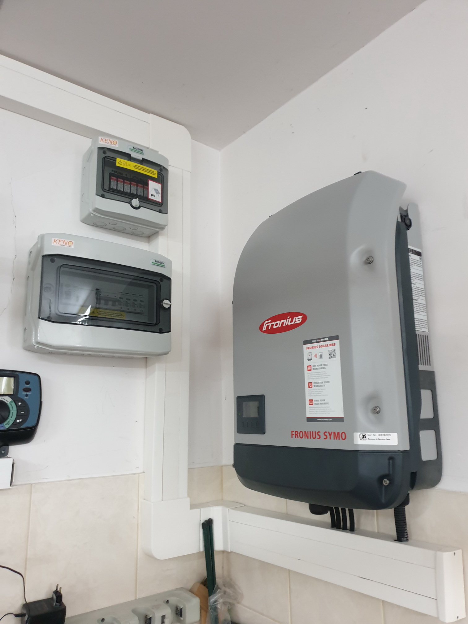Szary inwerter Fronius Symo zamontowany na ścianie obok dwóch szarych skrzynek KENO z zabezpieczeniami, instalacja fotowoltaiczna wewnątrz pomieszczenia.